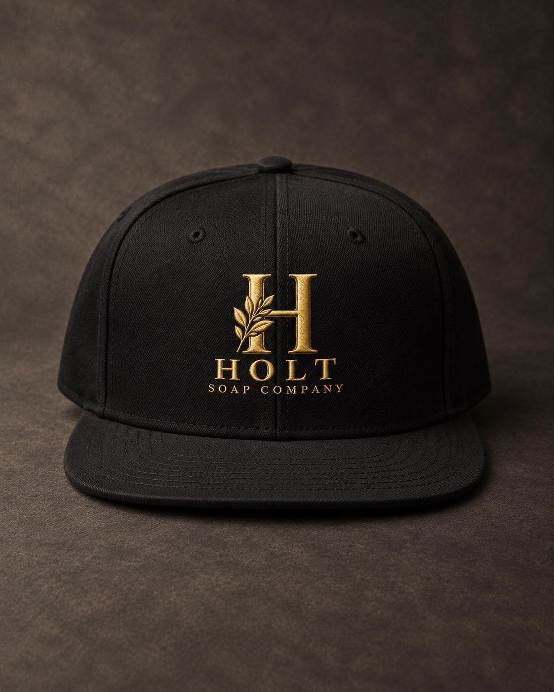 Black Holt flat-brim snapback with gold botanical monogram, resting on a charcoal surface